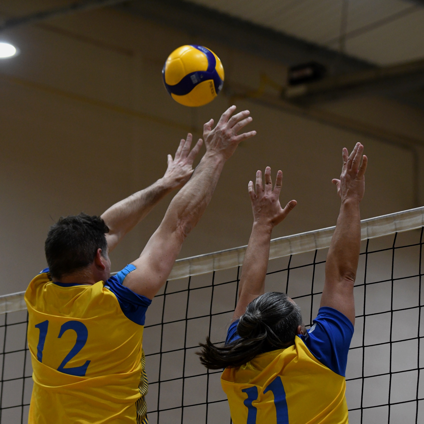 Two blockers in yellow rise above the net as the ball hangs overhead
